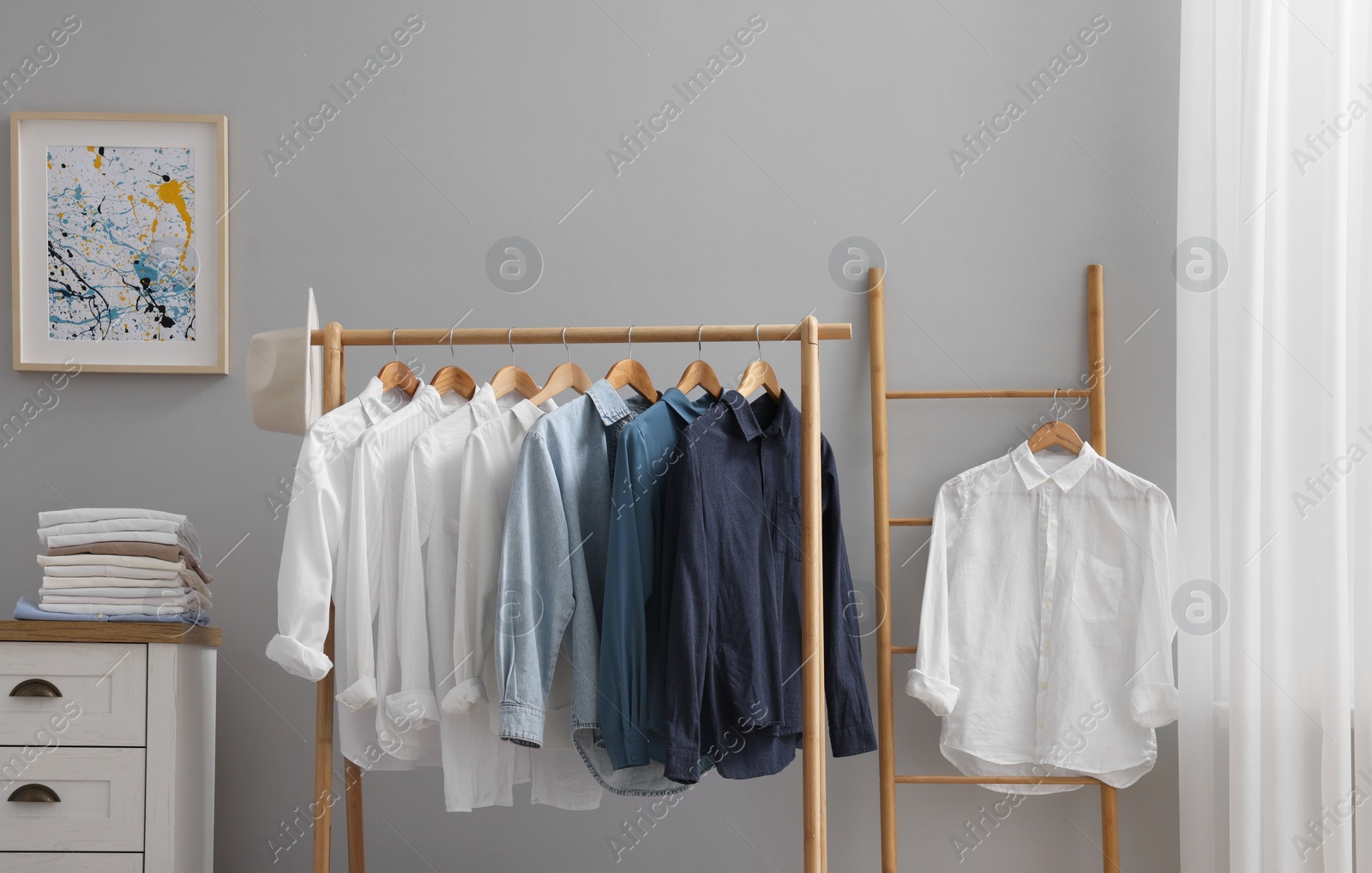 Photo of Wardrobe organization. Rack with different stylish clothes, hat and chest of drawers near grey wall in room