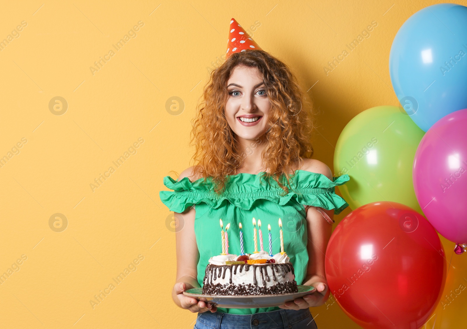 Photo of Young woman with birthday cake near bright balloons on color background