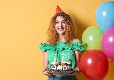 Young woman with birthday cake near bright balloons on color background