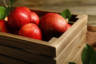 Ripe red apples with water drops in crate and green leaves on table, closeup