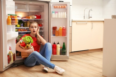 Photo of Emotional young woman sitting near refrigerator in kitchen