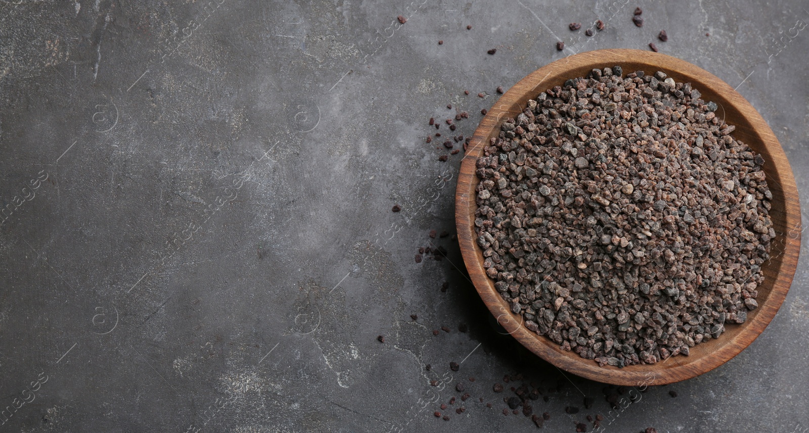 Photo of Black salt in wooden bowl on grey table, top view. Space for text