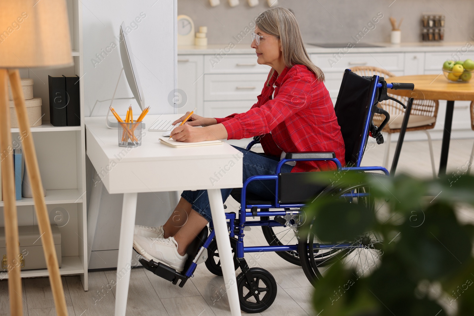 Photo of Woman in wheelchair writing in notebook while using computer at home