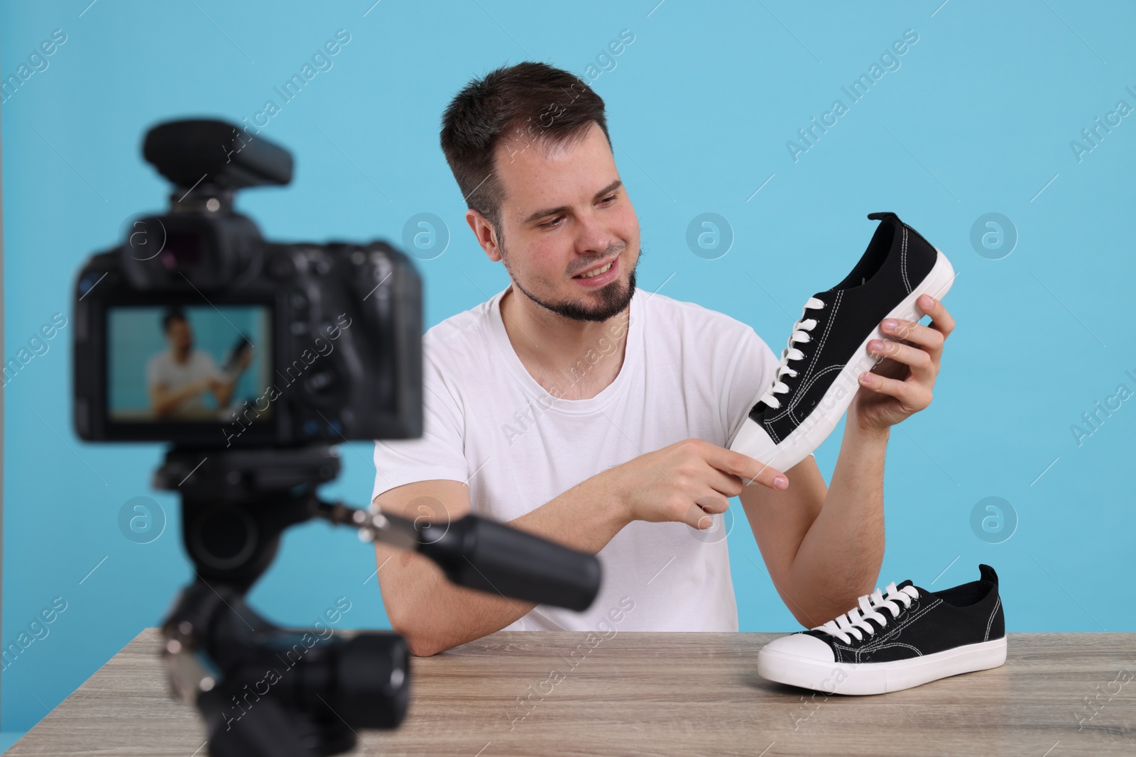 Photo of Smiling fashion blogger showing sneakers while recording video at table against light blue background