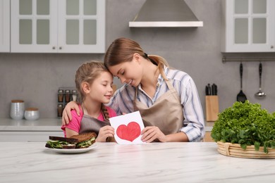 Photo of Little daughter congratulating mom with greeting card in kitchen. Happy Mother's Day