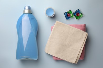 Photo of Bottle of fabric softener, laundry capsules and stacked clean clothes on light background, flat lay