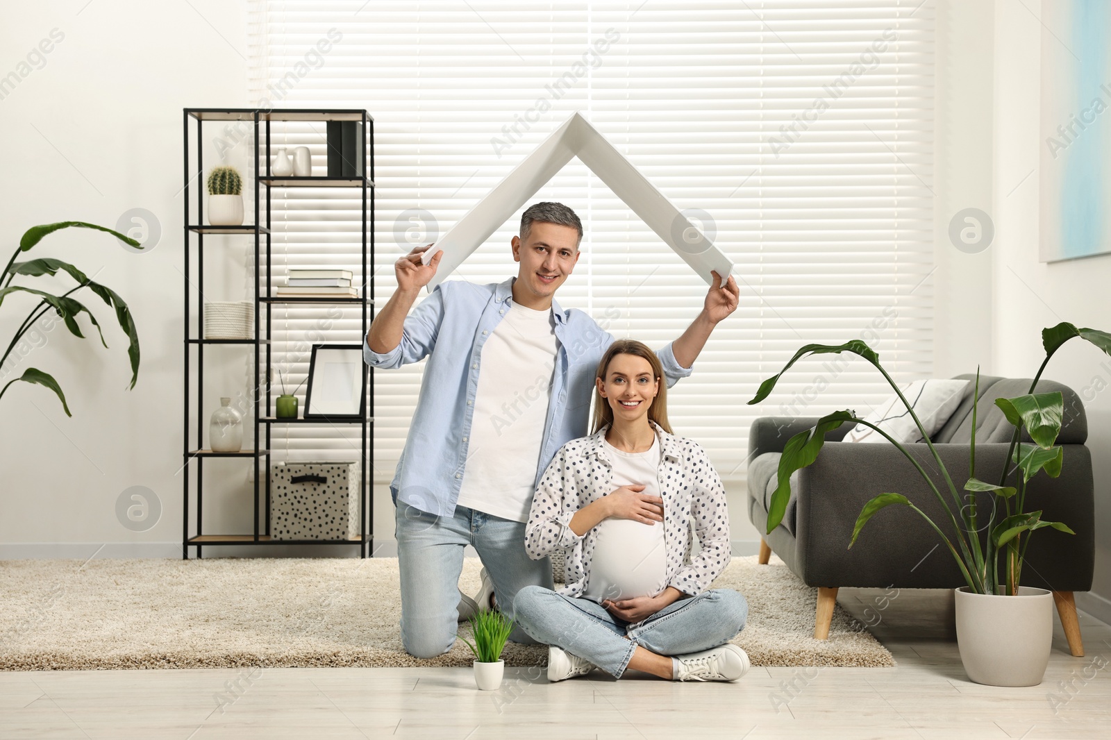 Photo of Young family housing concept. Man holding cardboard roof over his pregnant wife head at home