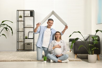 Young family housing concept. Man holding cardboard roof over his pregnant wife head at home