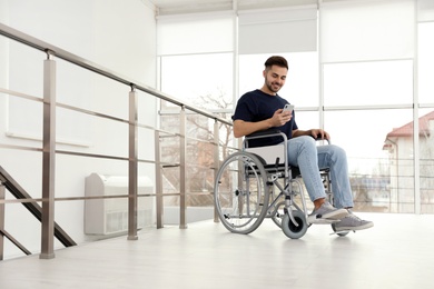 Photo of Young man in wheelchair using mobile phone near window indoors
