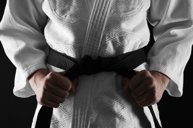 Photo of Man in keikogi tying black belt on dark background, closeup. Martial arts uniform