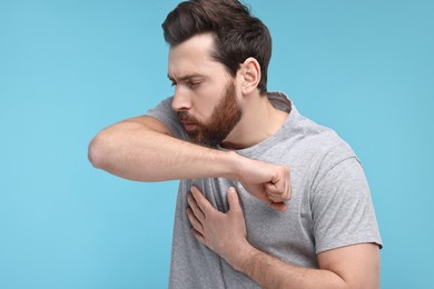 Sick man coughing into his elbow on light blue background. Cold symptoms