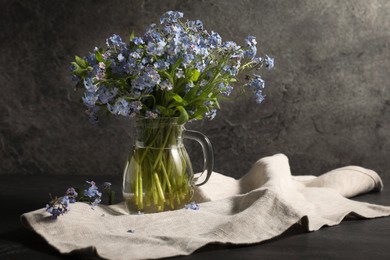 Bouquet of beautiful forget-me-not flowers in glass jug and cloth on gray table