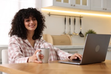 Beautiful young woman in stylish pyjama with cup of drink using laptop at wooden table in kitchen