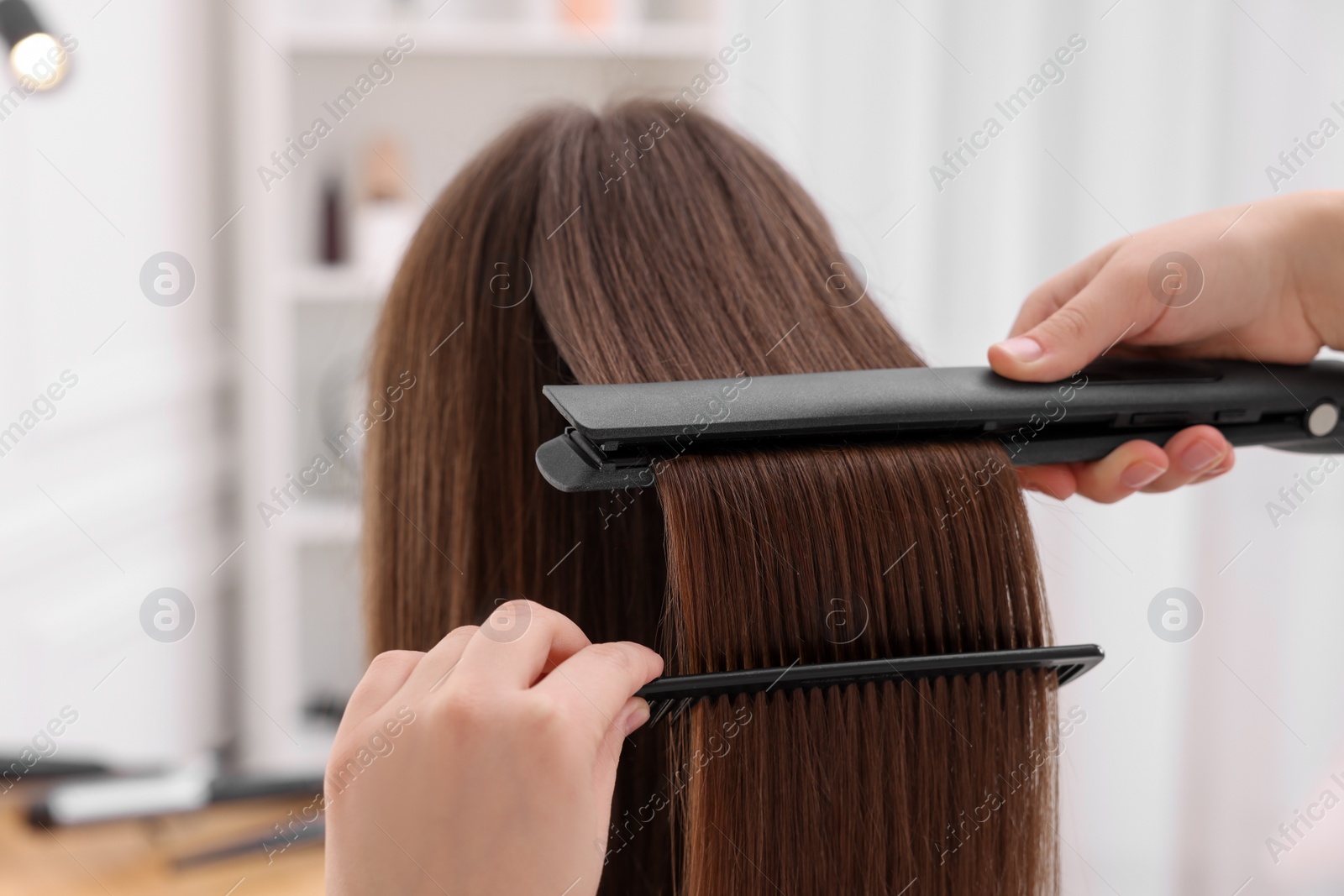 Photo of Hairdresser straightening woman's hair with flat iron in salon, closeup