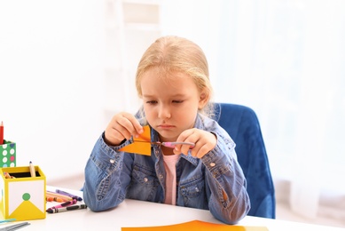 Upset little left-handed girl cutting orange color card with scissors at table
