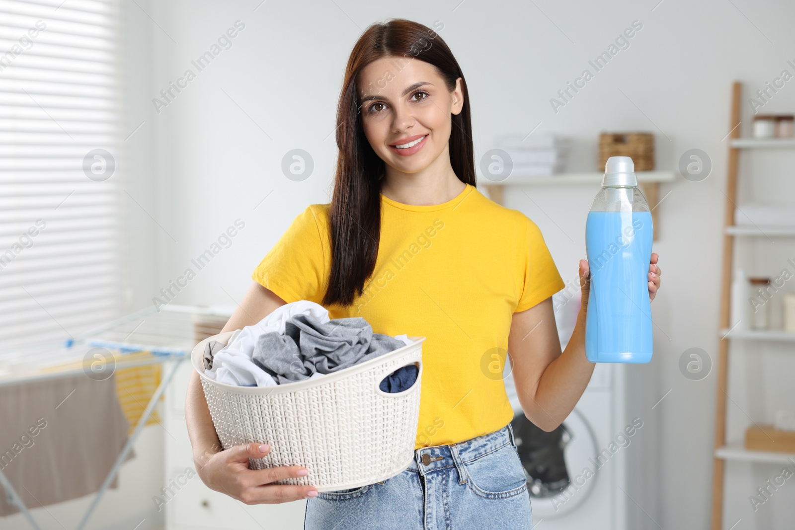 Photo of Woman holding basket with dirty clothes and fabric softener in bathroom