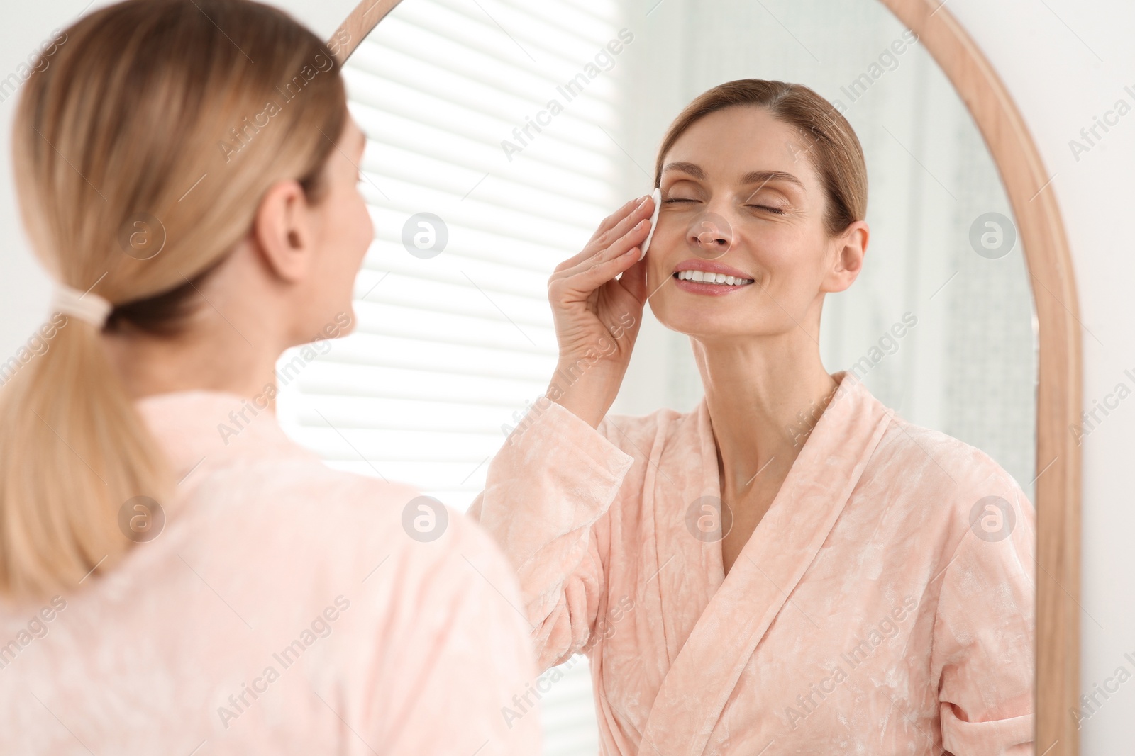 Photo of Beautiful woman removing makeup with cotton pad near mirror indoors