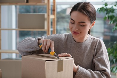 Photo of Young woman using utility knife to open parcel indoors