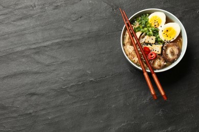 Photo of Noodle soup. Bowl of delicious ramen, ingredients and chopsticks on black table, top view. Space for text