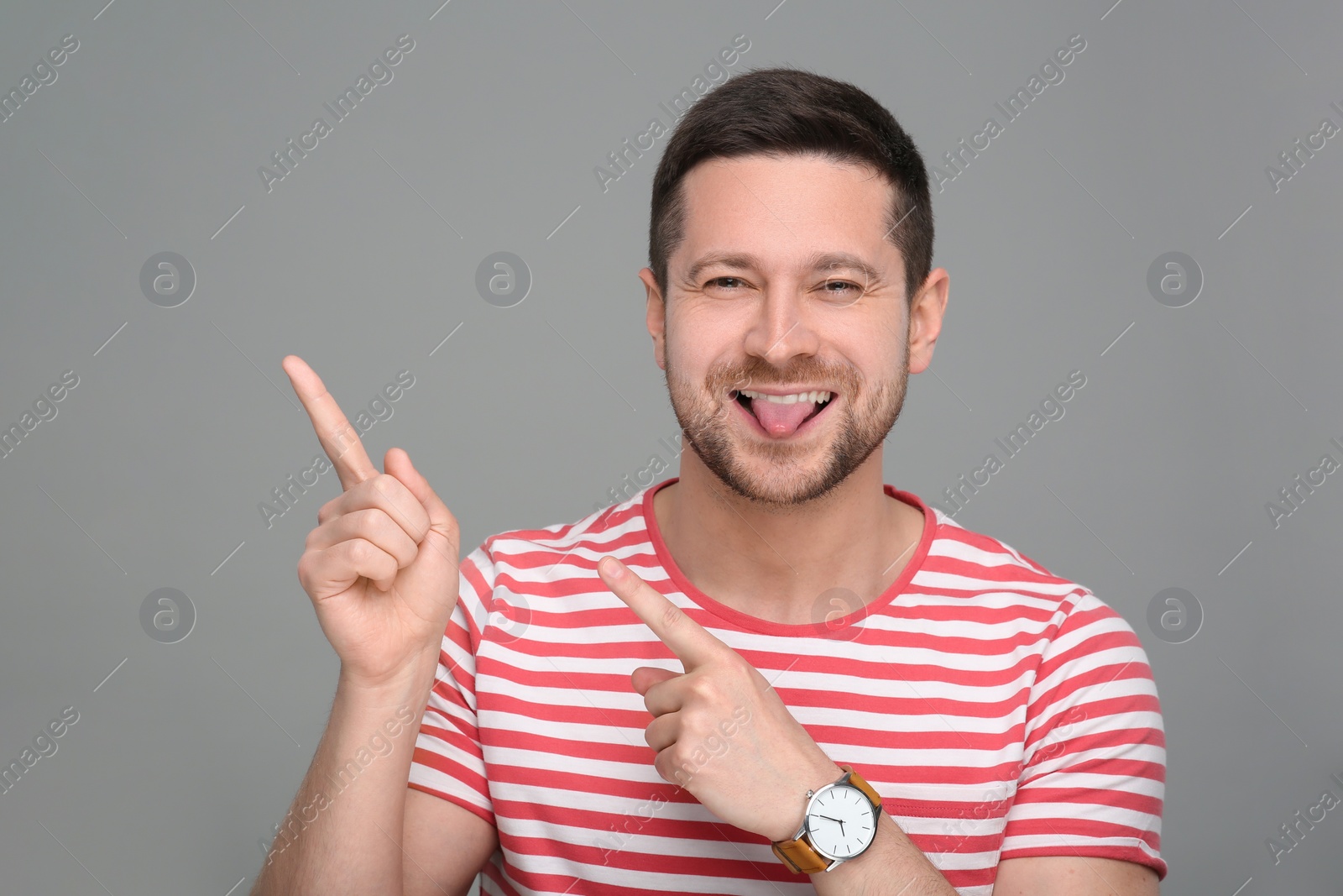 Photo of Happy man showing his tongue and pointing at something on grey background