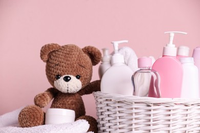 Photo of Baby cosmetic products in wicker basket, bath accessories and knitted toy bear on pink background