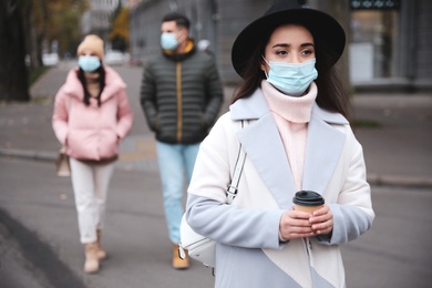 Photo of Young woman in medical face mask with cup of coffee walking outdoors. Personal protection during COVID-19 pandemic