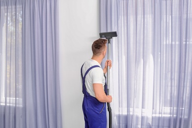 Photo of Male worker removing dust from curtains with professional vacuum cleaner indoors