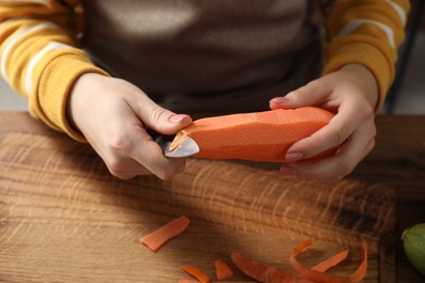 Photo of Woman peeling fresh carrot with knife at wooden table indoors, closeup