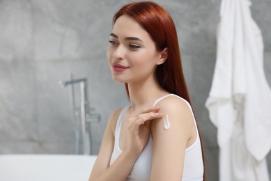 Photo of Beautiful young woman applying body cream onto shoulder in bathroom, space for text