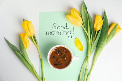 Photo of Aromatic coffee, beautiful flowers and card with words GOOD MORNING on white table, flat lay