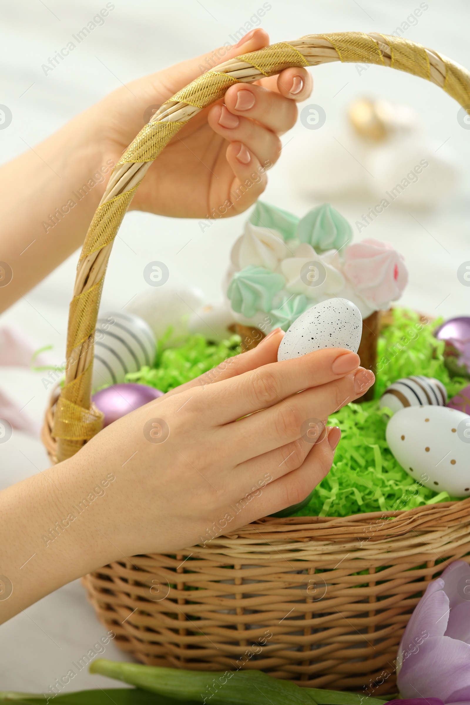 Photo of Woman putting painted egg into Easter basket at table, closeup
