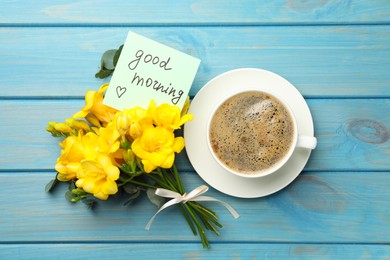 Photo of Cup of aromatic coffee, beautiful yellow freesias and Good Morning note on light blue wooden table, flat lay