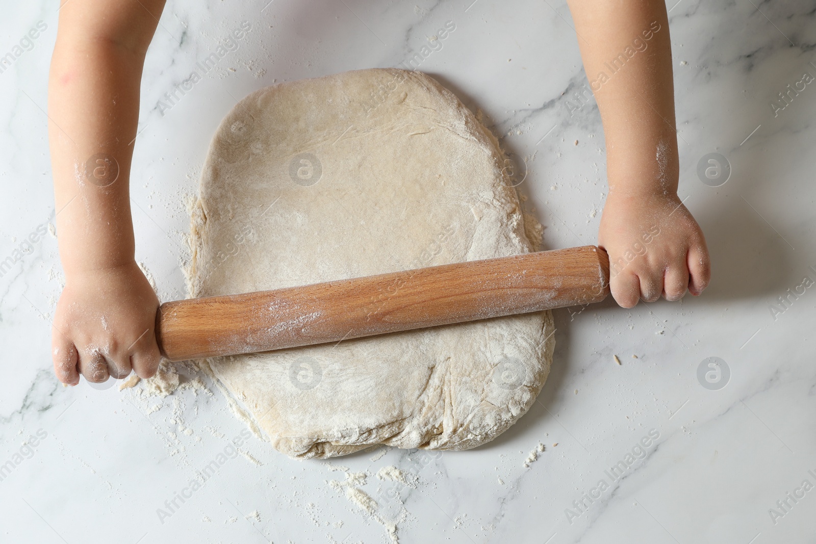 Photo of Little child rolling raw dough at white table, top view