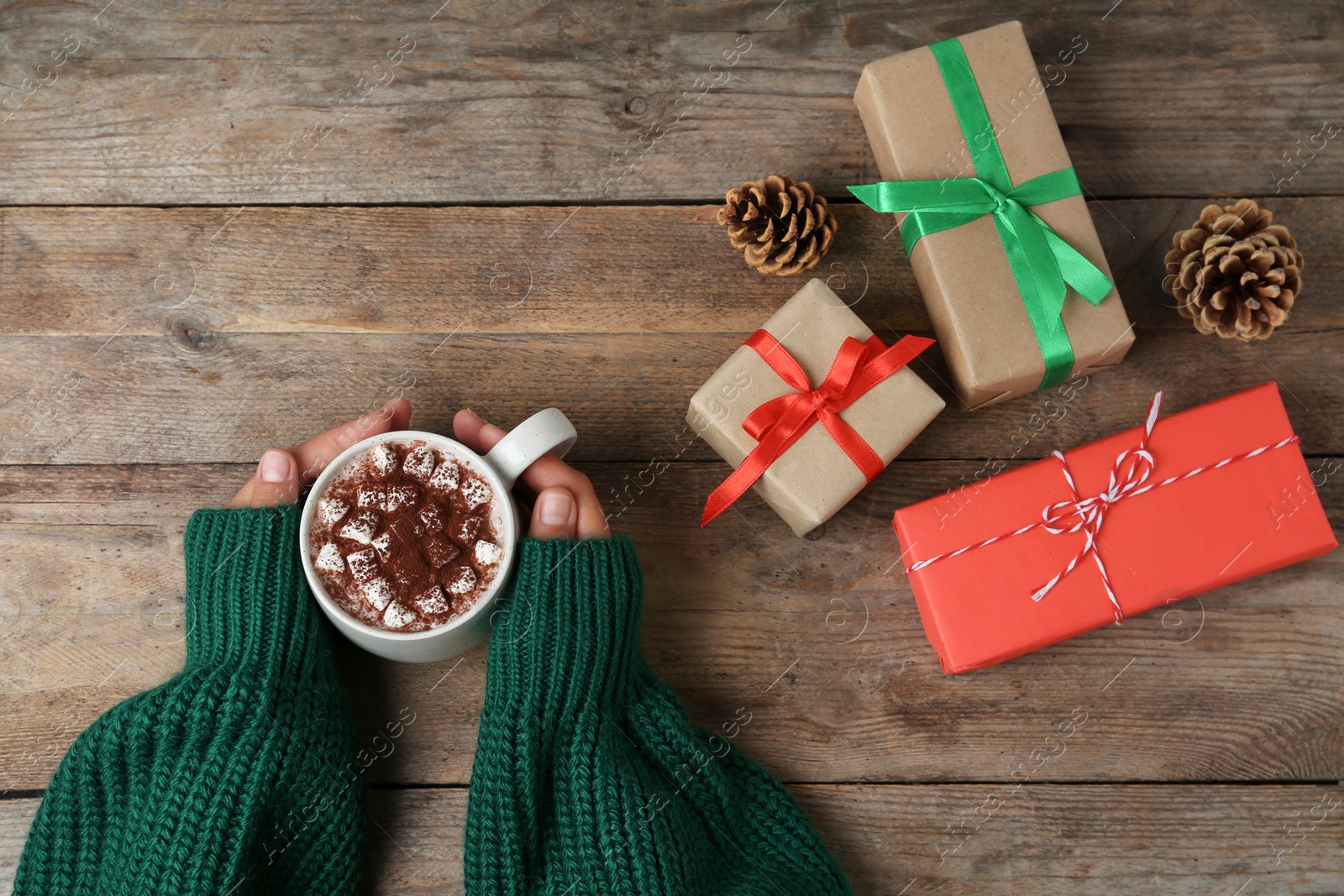 Photo of Woman holding cup of delicious cocoa drink at wooden table with Christmas presents, top view