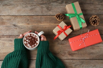 Photo of Woman holding cup of delicious cocoa drink at wooden table with Christmas presents, top view