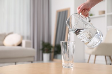 Photo of Woman pouring fresh water from jug into glass at table against blurred background, closeup. Space for text