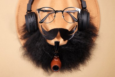 Man's face made of artificial mustache, beard, glasses and hat on beige background, top view