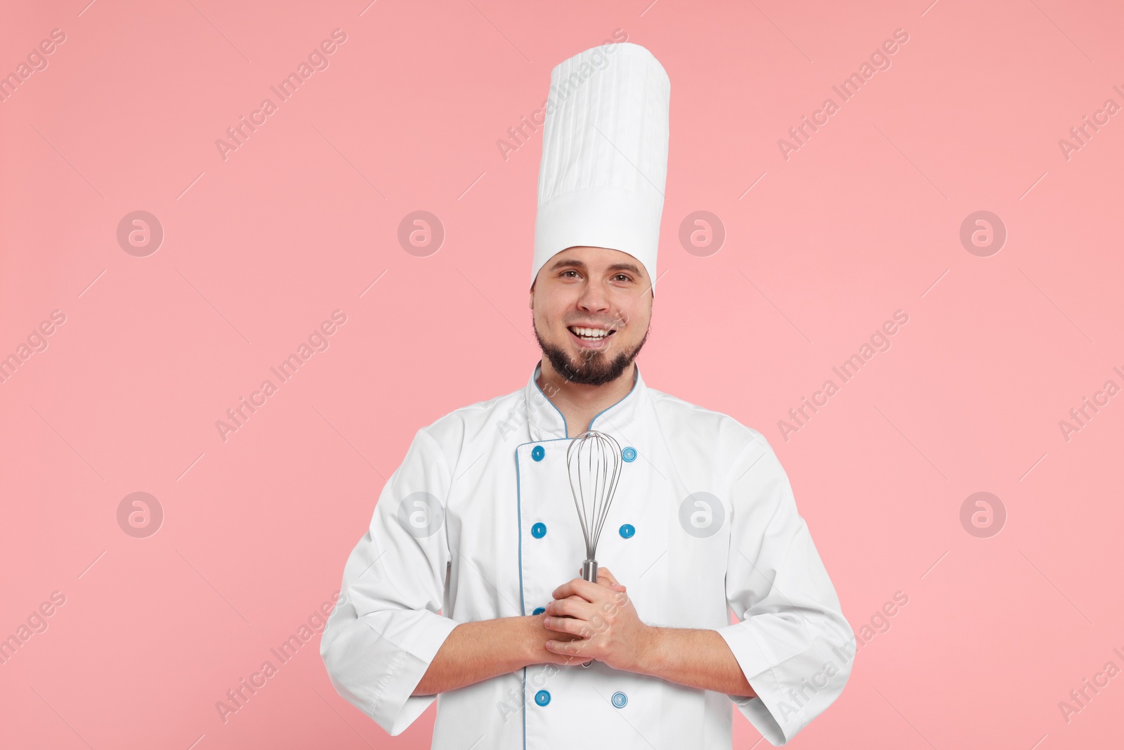 Photo of Happy professional confectioner in uniform holding whisk on pink background