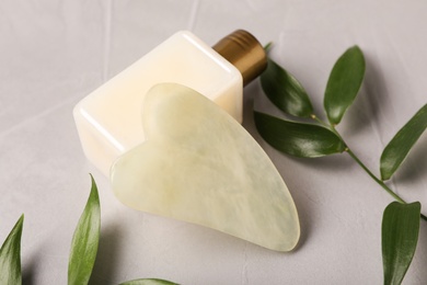 Jade gua sha tool, cosmetic product and green branches on grey table