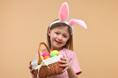 Photo of Easter celebration. Cute girl with bunny ears holding basket of painted eggs on beige background