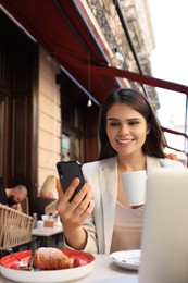 Photo of Beautiful young woman with cup of coffee using smartphone in outdoor cafe