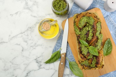 Photo of Freshly baked pesto bread with basil and knife on white marble table, flat lay. Space for text