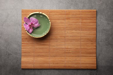 Photo of Bamboo mat and bowl with orchid flower on grey table, top view. Space for text