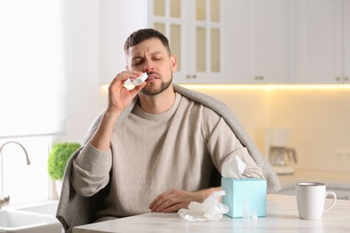 Photo of Ill man using nasal spray at table in kitchen