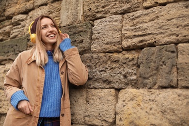 Photo of Young woman with headphones listening to music near stone wall. Space for text