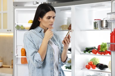 Thoughtful young woman with smartphone near modern refrigerator in kitchen