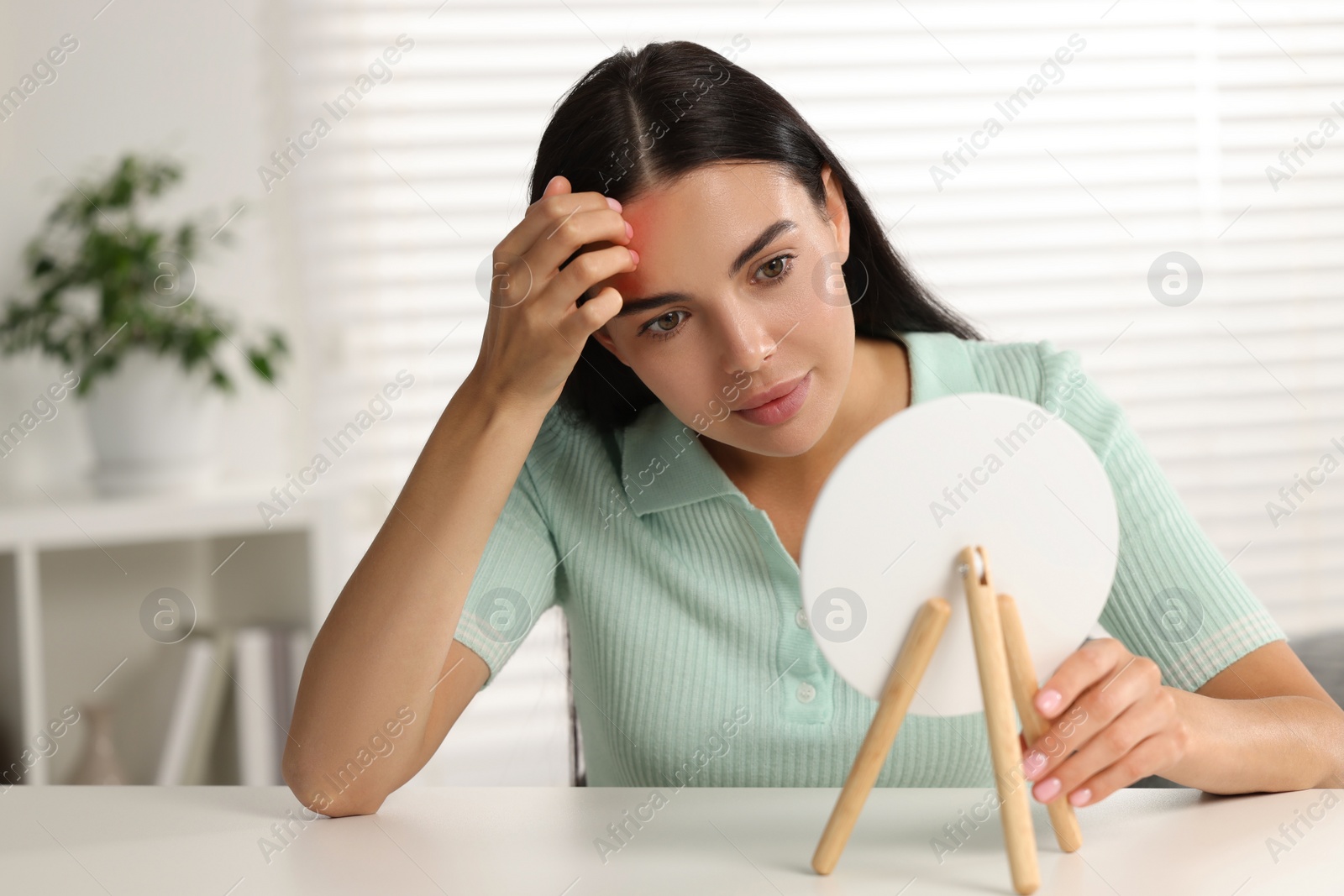 Photo of Suffering from allergy. Young woman looking in mirror and scratching her forehead at white table indoors