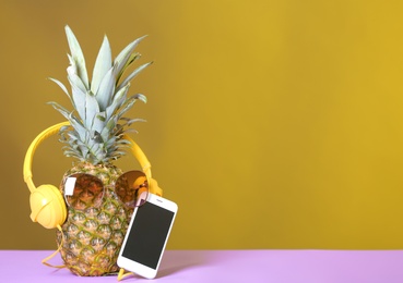 Photo of Pineapple with headphones, sunglasses and smartphone on table against color background. Space for text