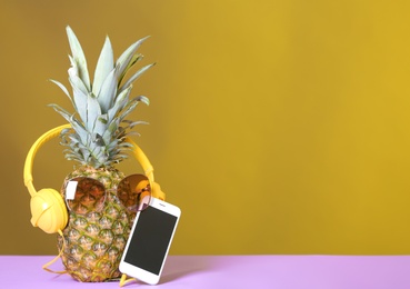 Pineapple with headphones, sunglasses and smartphone on table against color background. Space for text