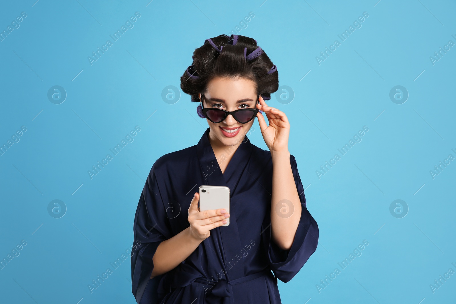 Photo of Happy young woman in silk bathrobe with hair curlers and sunglasses using smartphone on light blue background